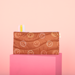 Light pink background with a brown, colorful smiley pattern puffy pouch with three jotter pens on a pink surface.