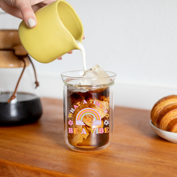 Creamer being poured from a ceramic dish into the iced coffee inside a "What A Time To Be A Vibe" glass tumbler.