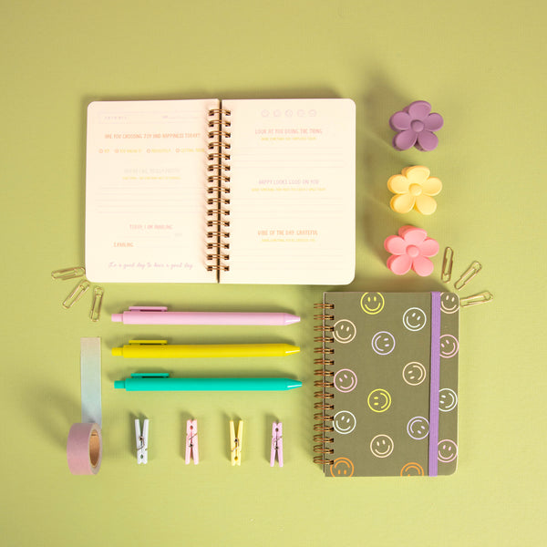 Green mini notebook with colorful smiley faces all around with purple strap and guided gratitude pages on a pastel green background with jotter pens and misc items