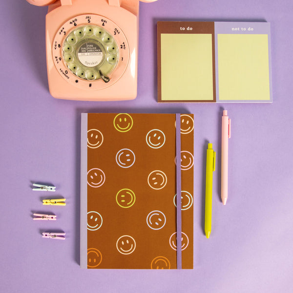 Brow smiley notebook with purple strap on a purple background with a tearaway and misc items.