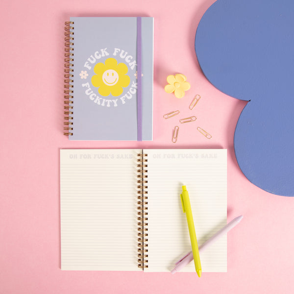 Pastel purple "fuck fuck fuckity fuck" notebook with a purple strap with colorfully lined paper and jotter pens with misc items on a pink background 