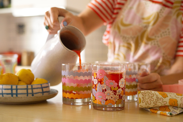 Someone pouring a drink into a scallop rocks glass cup while a gathering flowers glass cup has a drink inside of it.