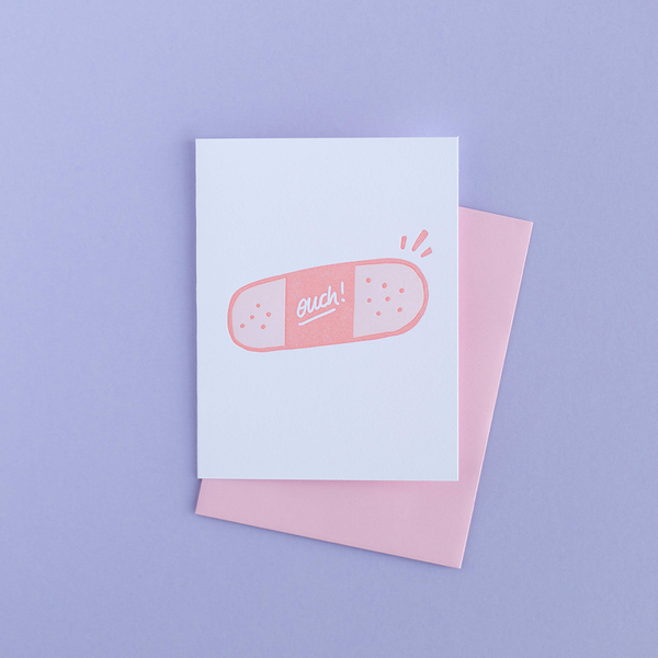 White greeting card with a medium pink and light pink bandaid with the text "ouch!" in white. There is a pink envelope and a purple background.