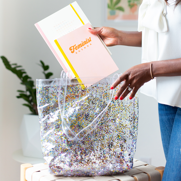 A clear vinyl tote bag filled with rainbow confetti sits on a stool. A woman is placing a notebook and a planner into the bag. The notebook on top says "Feminist Agenda". 