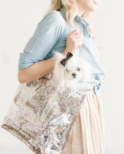 A woman stands holding a large confetti tote with a white small dog wearing a black bow. 