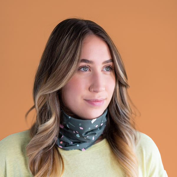 A multicolored terrazzo speckled mask-like item with a dark teal background color. Displayed on a person wearing it around their neck in front of an orange background.