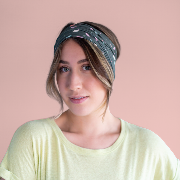 A multicolored terrazzo speckled mask-like item with a dark teal background color. Item is being used as a headband by a woman in front of a dusty pink background.