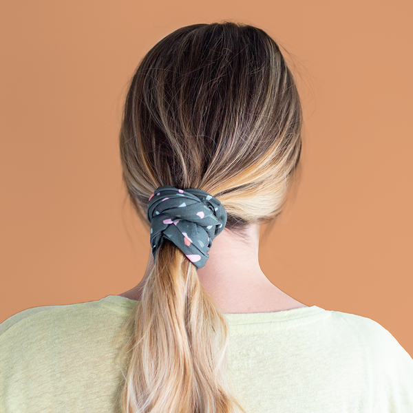 A multicolored terrazzo speckled mask-like item with a dark teal background color. Item is multifunctional and is being used as a hair tie. 