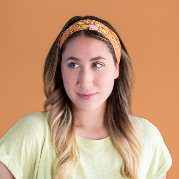 An orange, mask-like item, with multicolored and differently designed flowers that can be used as a headband. Displayed by person using neck-fluff as a headband.