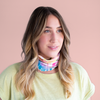 A rainbow tie-dye neck/face cover that is displayed on a woman's neck in front of a dusty pink background.