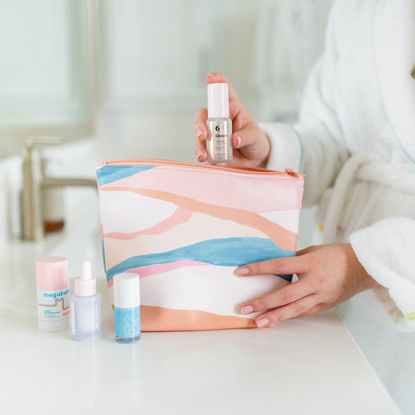 Lady in bathrobe placing self care products into a vegan leather light peach, blue, and pink pouch with a zip top.