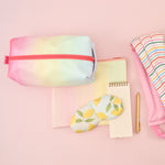 Meltdown Doppelganger is a rainbow print with a coral zipper and carrying handle. Displayed with and eyemask, a Daybreak Pouch, a To-do task pad, a Gold Jotter Pen, and a weighted Neck Wrap.