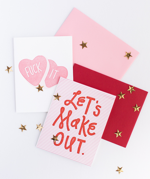 Image of two cards. The one on the left is white with pink hearts and the text "Fuck It". The one on the left is red and white striped with red text "Let's Make Out". Cards are sprinkled with gold star confetti.