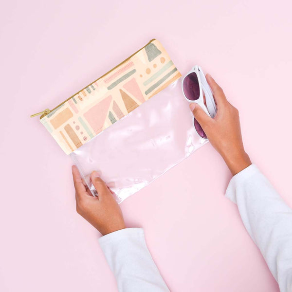 A clear vinyl pouch with the top of the bag having an abstract fruit design in cream colors and muted pastels like green and pink. Paint-like smears are also a part of the design. Pouch is being held by person with sunglasses in front of pink background.