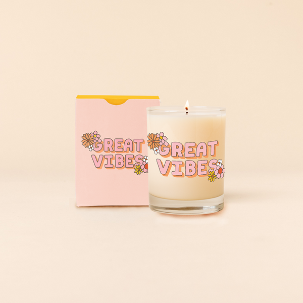 Rock glass candle reading "GREAT VIBES" with colorful cluster flower design at opposite corners of statement. Pastel pink box packaging with same design as candle. 