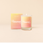 Candle rocks glass with yellow-to-orange ombre decal and text that reads "Taurus" with minimalist, white sparkle stars surrounding the text; "the one who holds your heart" sits at the bottom of the decal. Box packaging with the same design sits behind glass.