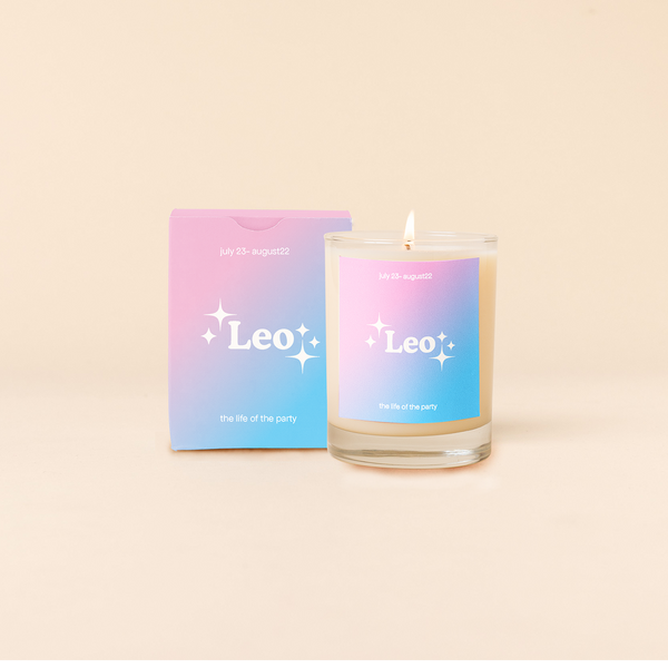 Candle rocks glass with pink-to-bright blue ombre decal and text that reads "Leo" with minimalist, white sparkle stars surrounding the text; "the life of the party" sits at the bottom of the decal. Box packaging with the same design sits behind glass.