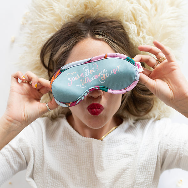 Girl laying on a fury white pillow with a jewel toned colorful sleep mask that says "You've got whatever it is".