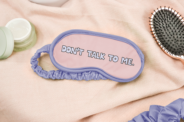 A sleep mask with periwinkle outline and strap that says don't talk to me laying next to a gold brush and moisturizer