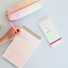 woman writing on a taskpad with a make it happen taskpad sitting next to it on a light blue background.