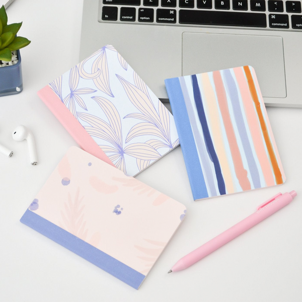 three mininotebooks next to a laptop, airpods, a plant, and a pink jotter pen. One notebook has periwinkle leaves, one has stripes, and one is a watercolor