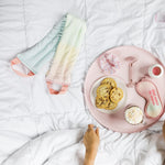 A bed setting with a woman reaching for self care items, a weighted neck wrap in orange to green ombre, a sleep mask, a clear vinyl pouch with lemons on the top with hair rollers inside.