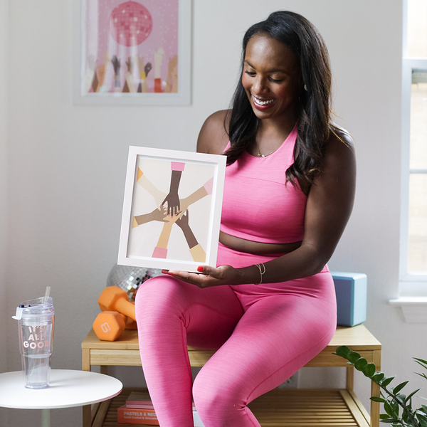 A woman holding a Join Hands Poster. The print is a design with hands of all skin types joining in and forming a circle-like formation in front of a peach background. The woman is holding the print in a home setting.