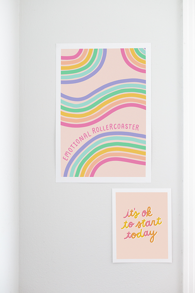 A pink poster with rainbow paths twisting back and forth, with "emotional rollercoaster" scripted in pink. Displayed on a white wall with another small prink hanging below the "Emotional Roller Coaster" print.