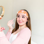 A woman wearing the Flower Power spa head wrap, holding a makeup brush near her eye and a mirror facing herself. The spa head wrap has a light pink trim, and a retro style flower pattern with an orangey-brown background. 