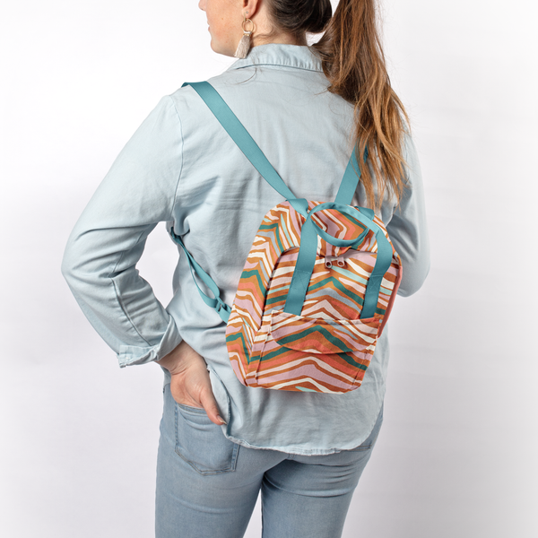 A multi-colored, animal-like printed mini backpack with a front pocket and bright blue shoulder straps. Displayed by person holding bag in front of a blank white background.