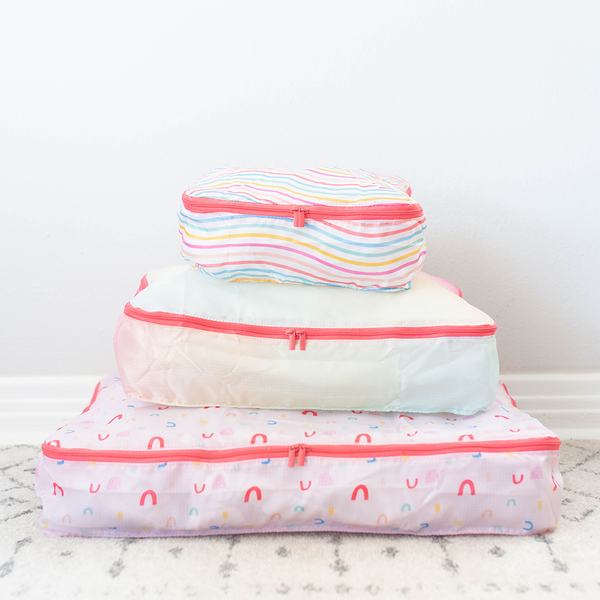 Image of 3 stacked packing cubes. One with multi colored wavy lines, one with a light rainbow gradient and one with multi colored rainbows turned different directions