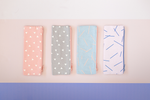 Four pouches sit on a surface. One with peach canvas material and white triangles. The second a grey background with white triangles. The third is a washed out denim with pink marks, and the fourth pouch is a light pink with navy marks.  