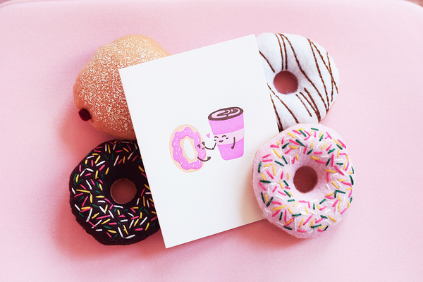 White greeting card with a cartoon donut and coffee cup holding hands. The card is surrounded by felt donuts. 