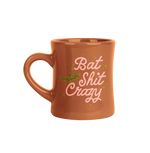 Camel color diner mug with "Bat Shit Crazy" written on the front in pink font, with illustration of bat and olive green minimalist sparkle-stars surrounding text.