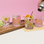 person pouring water into a set of 4 retro flower power rocks clear glass set sitting on a white table with a pink wall in the background.