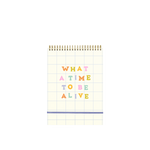 Taskpad with white grid design and text that reads "WHAT A TIME TO BE ALIVE" in multi-color letters on front, blue elastic enclosure at bottom and gold coil binding at top.