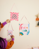 woman hanging up small flag with phrase festive festive festive in multi-color letters with pink background. next to it hanging is one large banner with the saying fest as fuck in multi-color letters with multi-color swirls around it and a medium banner with the phrase merry fucking christmas in green cursive font with pink and hot pink checkered pattern.