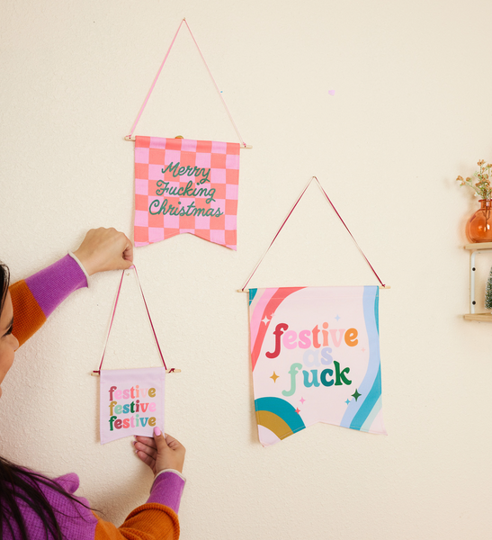 woman hanging up small flag with phrase festive festive festive in multi-color letters with pink background. next to it hanging is one large banner with the saying fest as fuck in multi-color letters with multi-color swirls around it and a medium banner with the phrase merry fucking christmas in green cursive font with pink and hot pink checkered pattern.