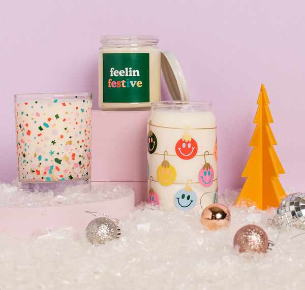 Three Holiday TOOT Candles, one Rocks Glass, one 12 oz. and one Yippie,being displayed with an orange acrylic tree decoration and small ornaments.