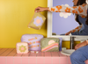 Mustard yellow background with a girl holding a medium waffle flower pouch on a pink square surface. All waffle pouches on the pink square surface along with misc skincare items. White mirror mirroring a purple and a white flower with a pink center background.