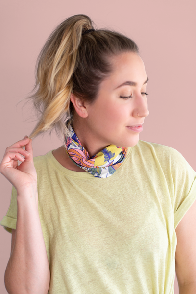 A floral collage neck/face covering with blues, oranges, yellows, and whites. Item is displayed around a persons neck