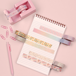 A notebook on a pink table with a pink tape dispenser, pens and paper clips. On the notebook six different washi take designs are taped onto the notebook page. 