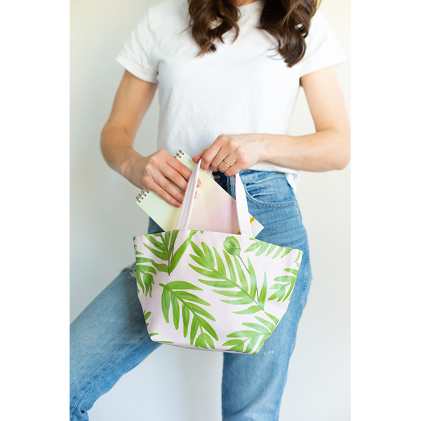 A woman holding a tote bag with flower buds and leaf sprigs all over in a blush pink base, she is pulling an ombre task-pad with top coil binding out of the bag.