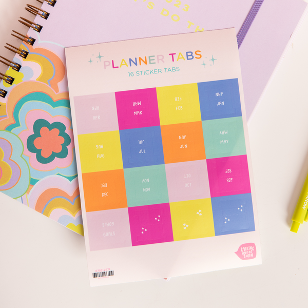 Sheet of planner tabs in "Trippy Dippy" color way, text with months of the year on each tab in white sitting on top of "Trippy Dippy" planner and surrounded by various stationery items.