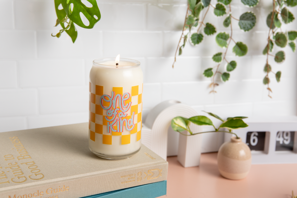 Can glass candle with yellow checkered print wrapped around glass; text that reads "one of a kind" on the front. Candle sits on books on a desk.