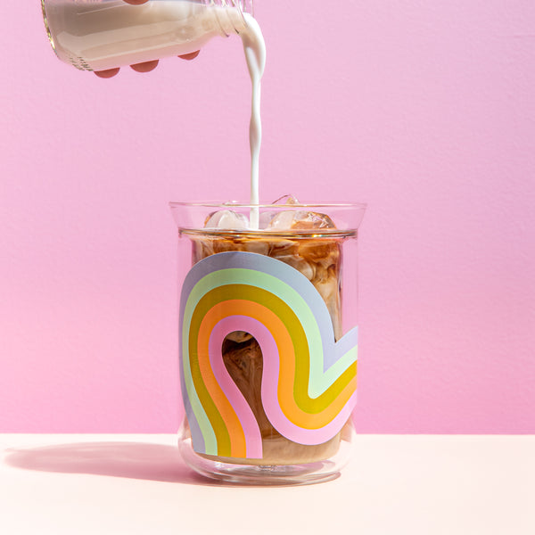 Multi-color waves element glass tumbler with coffee inside and someone adding milk. Pink background and white surface.