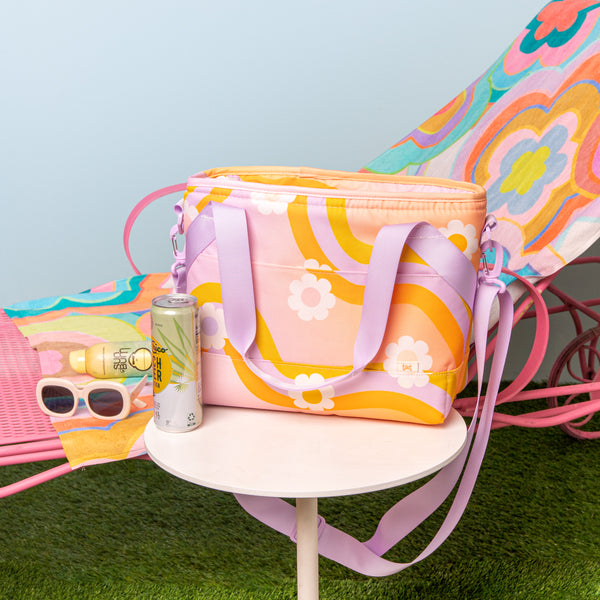 Trippy pattern beach towel with misc items on a beach lounge chair and a wavy daisy pattern ice princess bag on a white table. Light blue background.