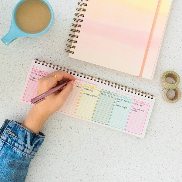 Hand filling out the colorful fresh start weekly deskpad next to her coffee.