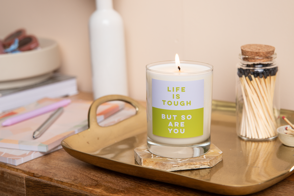 Candle rocks glass with blue and green label on front with text that reads "LIFE IS TOUGH, BUT SO ARE YOU". Candle sits on a gold tray surrounded by a jar of wooden matches and a variety of stationery.