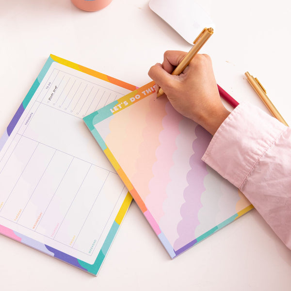 Girl writing on "let's do this" rising scallop tearaway notepad along with another tearaway notepad and gold jotter pen.